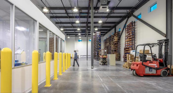 Warehouse with large shelves in the back and a room with windows on the left side next to large yellow pillars. There is an orange forklift on the right. 