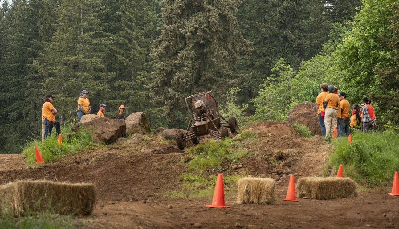 a team of people watching a baja sae vehicle drive in mud